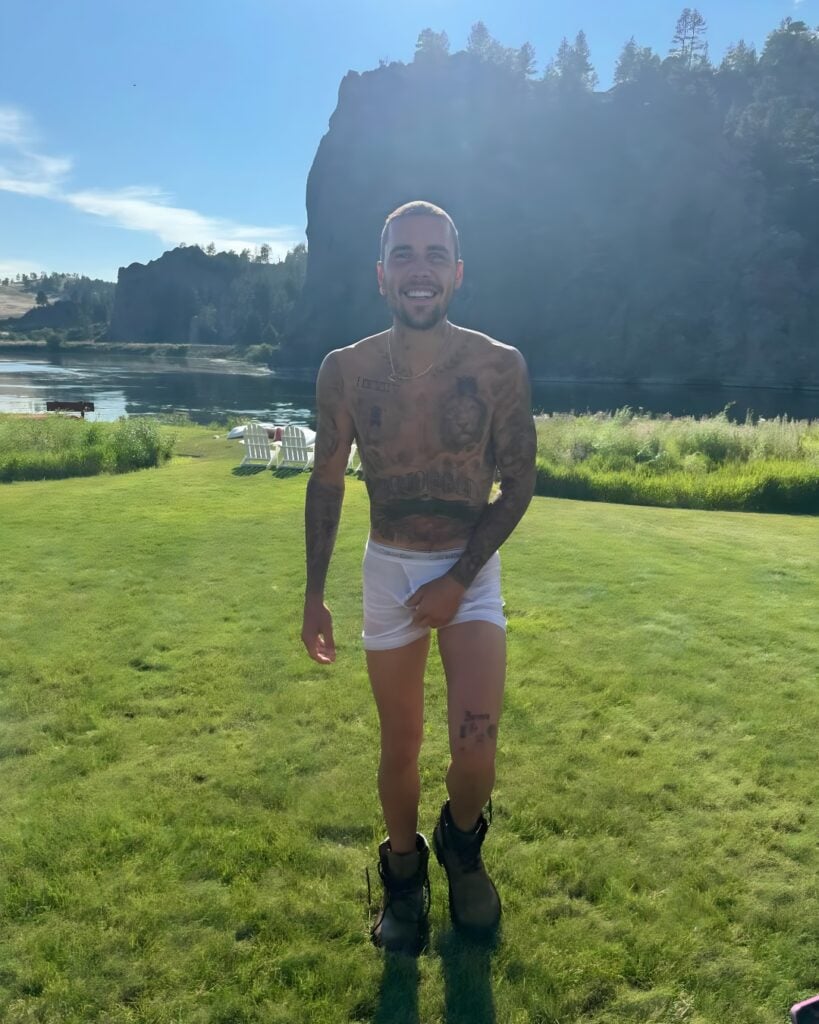 Justin Bieber mid-stride on a sunny lawn, wearing wet white boxer briefs and boots, smiling with a lake and cliff in the background.