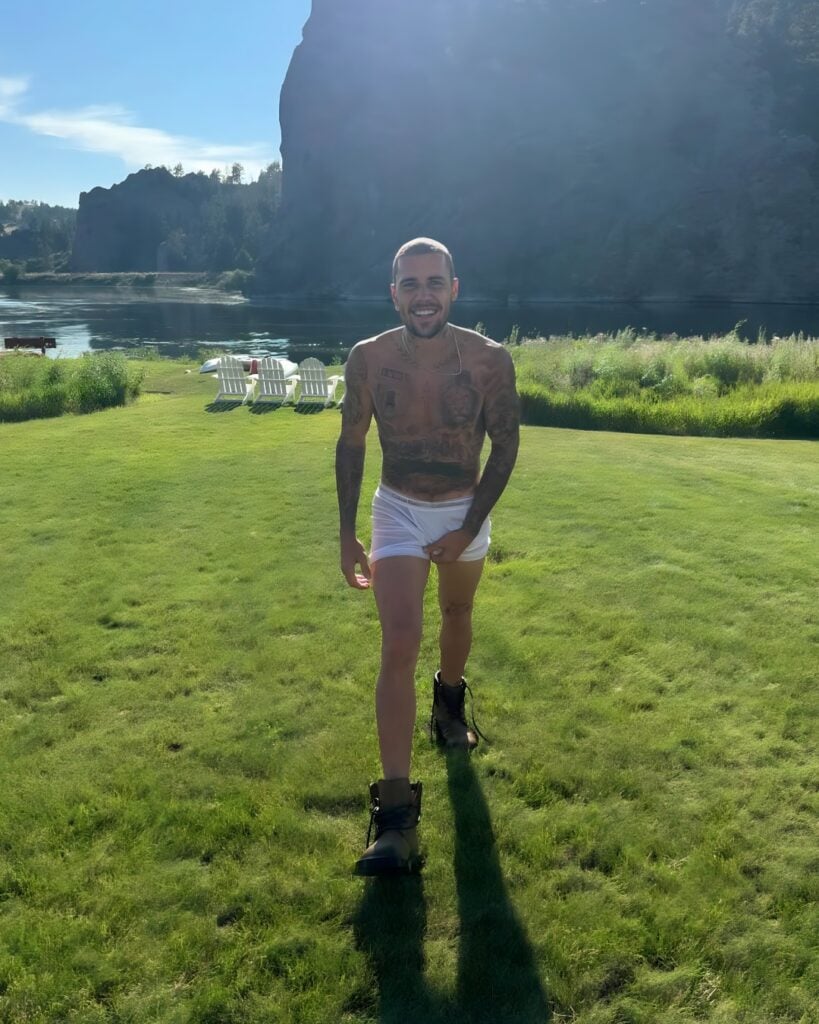 Justin Bieber walks toward the camera in soaked white boxer briefs and boots, smiling against a scenic lake and cliff backdrop.