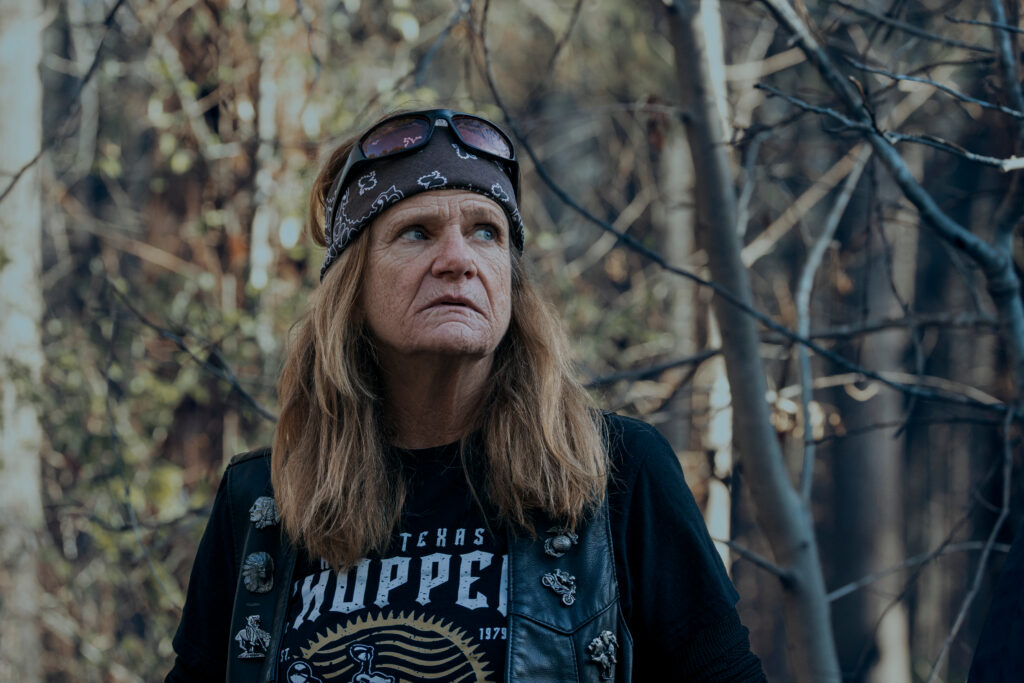 "Woman with long hair wearing a bandana, sunglasses, and biker vest stands outdoors in a forest, surrounded by trees and natural light, gazing off to the side with a serious expression."