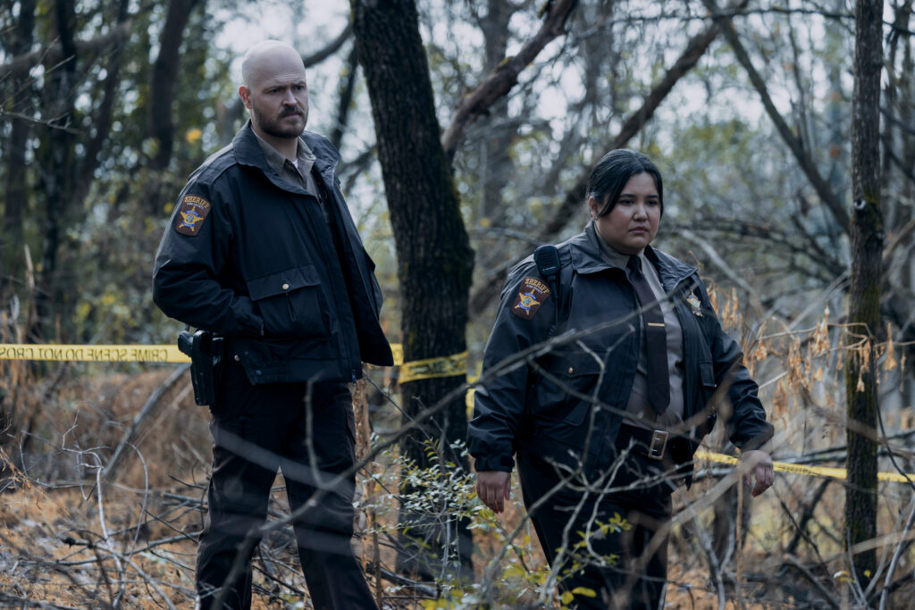 "Two uniformed police officers stand in a wooded area with yellow caution tape, appearing to investigate a scene amid trees and dry underbrush."