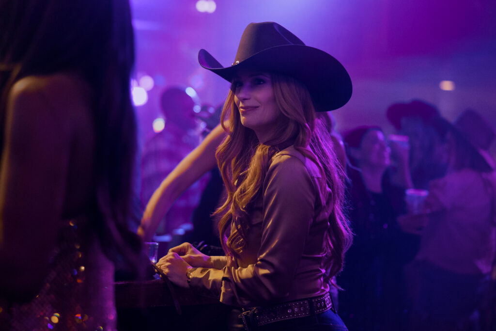 "Woman in a cowboy hat and western shirt stands by a bar in a dimly lit, lively nightclub or country bar with people in the background, colorful lights, and a festive atmosphere."