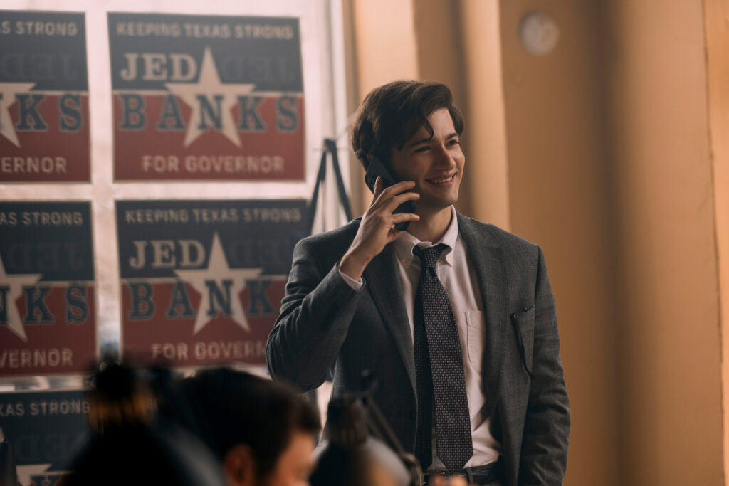 "Young man in a suit smiling while talking on a phone in an office decorated with political campaign posters that read "Jed Banks for Governor" and "Keeping Texas Strong." Warm indoor lighting with other people working in the background."