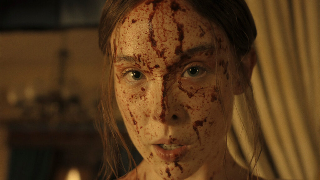 "A young woman with long hair, dressed in light clothing, has her face and upper chest covered in blood, standing indoors with dim warm lighting and blurred curtains and furnishings in the background."