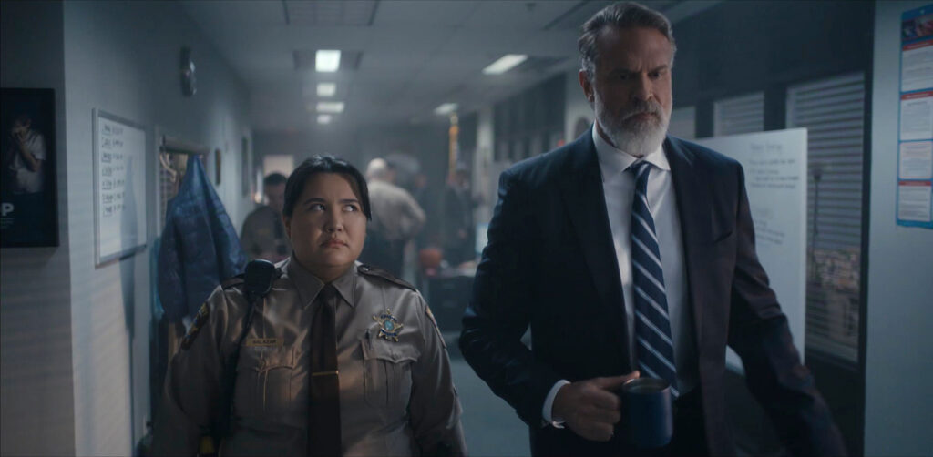 "A female sheriff and a man in a suit walk through a busy, fluorescent-lit police station hallway. The man holds a mug, and both appear serious, surrounded by officers and office equipment in the background."