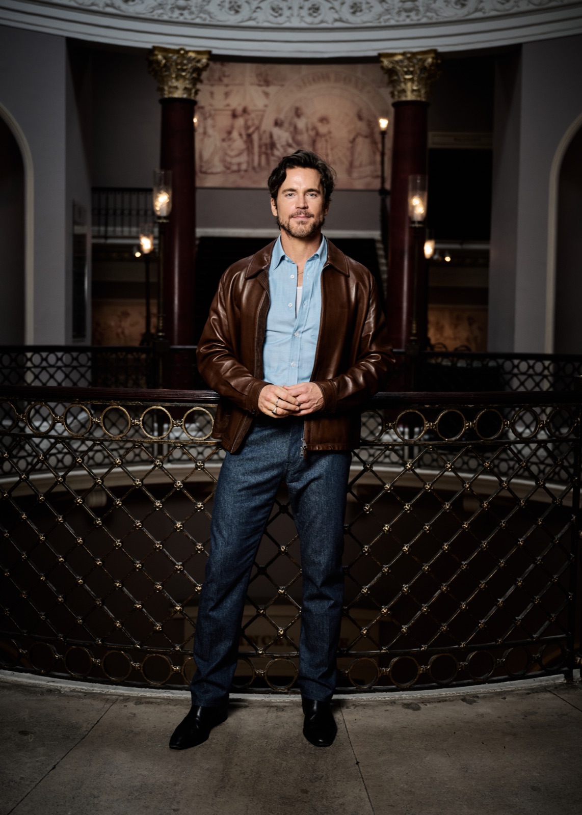 Matt Bomer wearing a brown leather jacket and blue shirt at Theatre Royal Drury Lane.